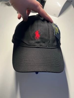 Ny Polo Ralph Lauren keps svart/röd – med tags – oanvänd - Stilren keps från Polo Ralph Lauren i klassisk svart design med röd broderad logga framtill. Helt ny och aldrig använd – tags sitter kvar. Perfekt till vardag, streetwear eller sportig outfit.  • Skick: Helt ny / oanvänd • Original tags kvar • Klassisk justerbar passform • Tidlös svart färg med röd logga • Perfekt present eller vardagskeps  polo keps, ralph lauren cap, svart keps, baseball cap, streetwear keps, justerbar keps, unisex keps, polo sport, klassisk keps