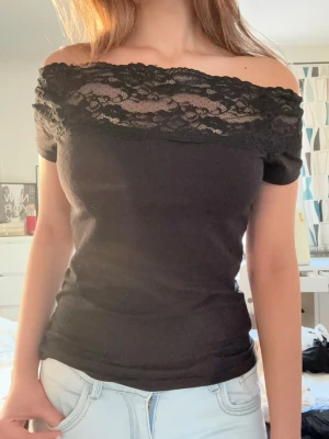 Svart offshoulder topp med spets - Snygg svart offshoulder topp med spetsdetaljer upptill. Toppen är tight och har korta ärmar. Perfekt för dig som vill ha en stilren men ändå lite extra look. Materialet känns mjukt och stretchigt, troligen bomull eller polyester.