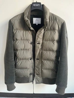Olivgrön cardigan från Woolrich - Olivgrön jacka från Woolrich med quiltad front i nylon och stickade ärmar i ull. Jackan har ribbad krage, muddar och nederkant samt stängs med både dragkedja och knappar. Nypris ca 5000kr. Skick 10/10. Hör av dig vid funderingar!
