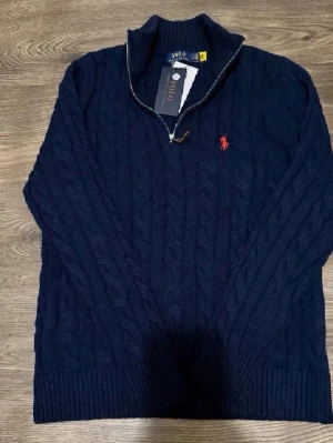 Mörkblå kabelstickad tröja Polo Ralph Lauren - Mörkblå kabelstickad tröja från Polo Ralph Lauren med halv dragkedja och röd broderad logga på bröstet. Tröjan har hög krage, ribbstickade muddar och är tillverkad i ett mjukt, värmande material. Perfekt för lager-på-lager-stil.