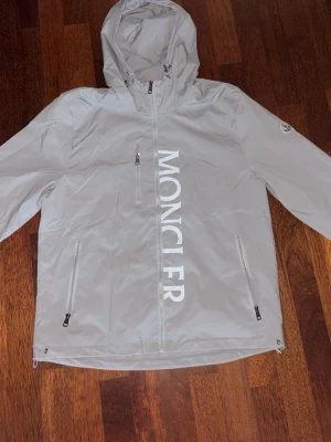 Grå vindjacka från Moncler - Ljusgrå Moncler-jacka med huva och dragkedja fram. Har Moncler-logotypen på ärmen och stor text framtill.  ⭐️ Skick: 9/10 🎨 Färg: Ljusgrå 📦 Snabb och noggrann leverans
