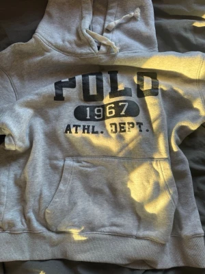 Grå hoodie från Polo Ralph Lauren - Grå hoodie från Polo Ralph Lauren med svart tryck 'POLO 1967 ATHL. DEPT.' på bröstet. Klassisk känguruficka framtill och justerbar huva med dragsko. Perfekt för en avslappnad och sportig stil.