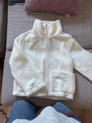 Vit teddyjacka med dragkedja - Mysig vit teddyjacka i fluffigt material med hög krage och dragkedja framtill. Jackan har två stora fickor och en avslappnad passform. Perfekt för kalla dagar när du vill vara både varm och stilren.