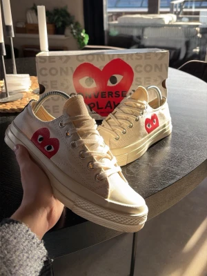 Converse x Comme des Garçons Play sneakers - Säljer ett par Converse x Comme des Garçons Play sneakers i offwhite canvas med den ikoniska röda hjärtloggan på sidan. Skorna har klassisk låg modell, vita gummisulor och silverfärgade metallöljetter. Perfekt för dig som vill ha något unikt och trendigt.