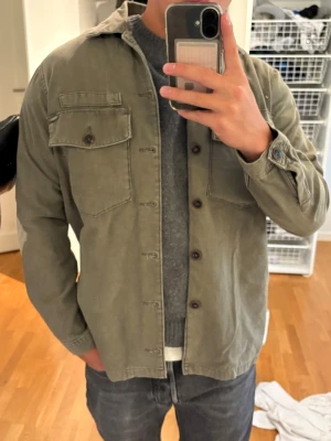 Grön overshirt - Säljer en olivgrön overshirt med klassisk krage och knappar framtill. Två bröstfickor med knappstängning ger en cool och avslappnad look. Perfekt att slänga över en t-shirt eller tröja för extra lager.
