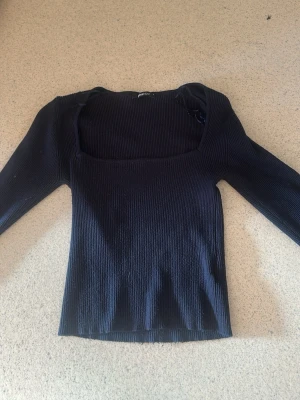 Svart ribbstickad topp Gina Tricot - Säljer en mörk blå ribbstickad topp från Gina Tricot med fyrkantig halsringning och långa ärmar. Materialet är mjukt och stretchigt, perfekt för att skapa en snygg och enkel look. Passar till många olika outfits.