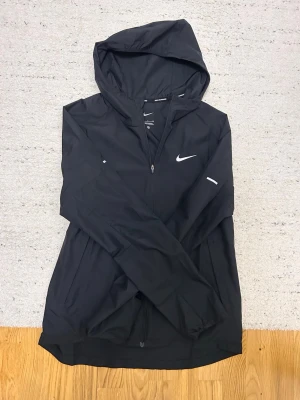 Svart Nike vindjacka med huva - Svart vindjacka från Nike med huva och hel dragkedja framtill. Jackan har två sidofickor med dragkedja och vita reflexdetaljer på ärmarna samt Nike-logga på bröstet. Tillverkad i lätt och smidigt material, perfekt för blåsiga dagar.