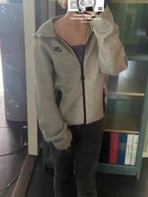 Grå zip-hoodie från Nike - Grå hoodie med dragkedja från Nike. Klassisk modell med svart Nike-logga på bröstet och svarta detaljer vid fickorna. Mjuk och bekväm, perfekt för en avslappnad stil. Huva och långa ärmar. Tillverkad i bomullsblandning.