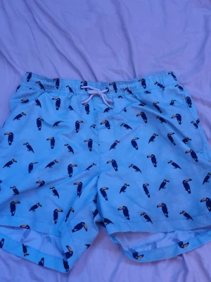 Blå badbyxor med tukaner Stock & Hank - Snygga ljusblå badbyxor från Stock & Hank med ett lekfullt mönster av tukaner. Shortsen har vit snörning i midjan och innerbyxa i mesh för extra komfort. Perfekta för strand eller poolhäng!
