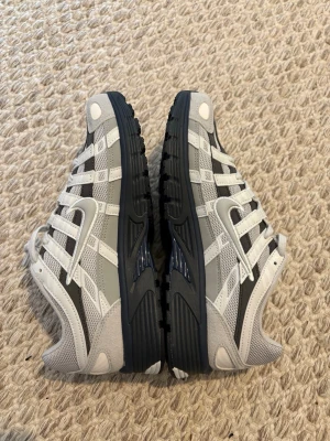 Nike grå och vita sneakers - Snygga sneakers från Nike i grått och vitt med mesh och mockadetaljer. Skorna har en sportig design med flera lager och tydliga ränder längs sidorna samt den klassiska swoosh-loggan. Yttersulan är svart och ger bra grepp. Perfekta för en streetwear-look.