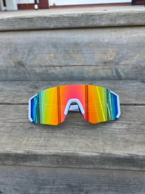 Sportiga regnbågsfärgade solglasögon - One size. Unisex. Nya. Säljer ett par riktigt snygga sportiga solglasögon med spegelglas i regnbågsfärger och vit ram. De har en futuristisk, kantig form och passar perfekt för dig som vill sticka ut. Perfekta för cykling, löpning eller bara för att se cool ut i solen.