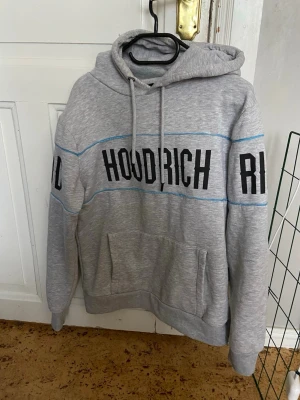 Grå hoodie från Hoodrich med tryck - Snygg grå hoodie från Hoodrich med svart tryck över bröstet och armarna. Hoodien har huva med dragsko, känguruficka framtill och blå detaljerade sömmar. Tillverkad i mjukt material som känns skönt mot huden. Perfekt för en avslappnad streetwear-look.