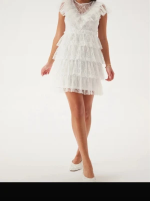 Student klänning  - Kort vit klänning med flera lager volanger och spetsdetaljer. Klänningen har v-ringning, transparenta partier upptill och är ärmlös. Perfekt för dig som vill ha en romantisk och lekfull look. Perfekt till studenten!