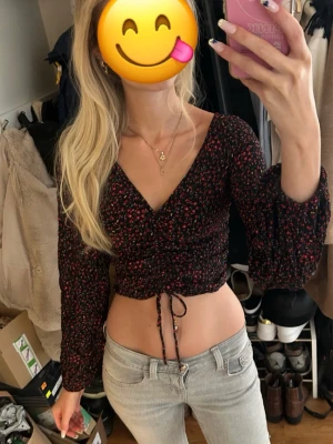 Blommig croppad topp - Trendig croppad topp med blommigt mönster i rött och grönt på svart botten. Toppen har långa ärmar, v-ringning och snörning i midjan för justerbar passform. Perfekt för dig som vill ha en feminin och cool stil. Ingen lapp för storlek finns. Skriv för exakta mått. 