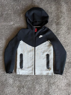 Nike Tech Fleece hoodie svart/grå - Nike Tech Fleece hoodie i svart och ljusgrått med dragkedja framtill. Hoodien har två fickor med svarta dragkedjor och en vit Nike-logga på bröstet. Tillverkad i mjukt och stretchigt material, perfekt för en sportig och avslappnad stil.