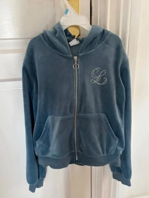 Blå velour hoodie med strassdetalj - Mysig blå hoodie i mjuk velour från Lindex. Dragkedja framtill med rund metallring, två stora fickor och huva. Bröstet pryds av ett L i strass. Perfekt för en chill och trendig look.