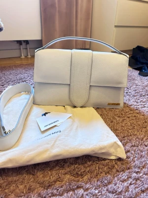 Le Grand Bambino ljusgrå handväska från Jacquemus - Snygg vit handväska från Jacquemus i stilren, rektangulär modell med guldfärgad logga. Väskan är gjord i läder och har både handtag och avtagbar axelrem. Perfekt för dig som gillar minimalistisk design och vill ha en trendig accessoar.