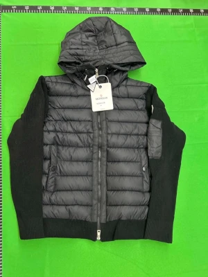 Cardigan  - Snygg svart pufferjacka från Moncler med vadderad front och stickade ärmar. Jackan har en stor huva, dragkedja framtill, fickor med knapp och en extra ficka på ärmen. Perfekt för kalla vinterdagar och riktigt trendig look.