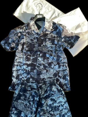 Blommigt set från Louis Vuitton - Säljer ett snyggt set från Louis Vuitton med kortärmad skjorta och matchande shorts. Setet har ett blått och vitt blommönster och Louis Vuitton-logga broderad på bröstet och shortsen. Materialet ser ut som denim eller bomullsmix. Perfekt för dig som vill sticka ut!