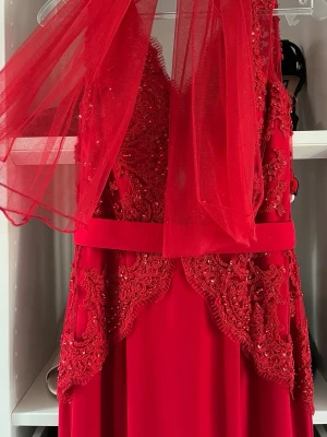 Röd långklänning - Bal  - Elegant röd långklänning från Rever Design med spetsdetaljer, pärlor och broderier. Klänningen har ett matchande rött bälte i midjan och ett lager av tunn tyll upptill (man kan använda tyllen som en sjal). Perfekt för dig som vill sticka ut med en glamorös look. NYPRIS 2500
