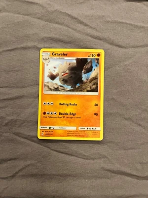Pokémonkort Graveler - Säljer ett Pokémonkort med Graveler, en orange och gul design med bild på Graveler i mitten. Kortet är tillverkat av papper och har attackerna Rolling Rocks och Double-Edge. Perfekt för samlare eller Pokémon-fans.