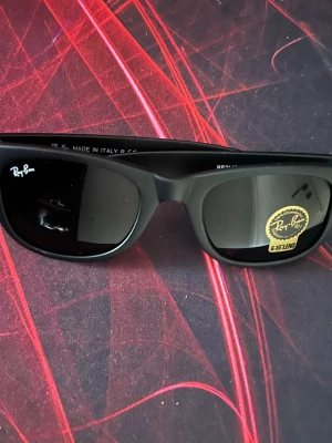 Svarta Ray-Ban solglasögon - Ray-Ban solglasögon med klassisk svart båge och mörka linser. Modellen har en tidlös och cool design med Ray-Ban-logga på både lins och skalmar. Perfekt för dig som vill ha en ikonisk look och skydda ögonen mot solen.