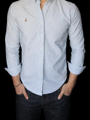 Randig skjorta från Ralph Lauren - Klassisk blå- och vitrandig skjorta från Ralph Lauren med button-down krage och broderad logga på bröstet. Skjortan har långa ärmar och knäppning framtill. Perfekt för en stilren och tidlös look.