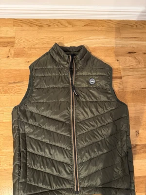 Olivgrön dunväst från Jack & Jones - Olivgrön dunväst från Jack & Jones med snygga chevron-sömmar och hög krage. Västen har en svart dragkedja med kontrastdetaljer och ett rundat märke på bröstet. Perfekt för lager-på-lager och till jeans eller hoodie.