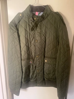 Olivgrön quiltad Barbour jacka XXL - Säljer en olivgrön quiltad jacka från Barbour International, Stone The Crows Collection. Jackan har ribbade muddar, två fickor med tryckknappar och en cool detalj med spänne vid kragen. Perfekt för dig som gillar klassisk brittisk stil och vill ha något som sticker ut.