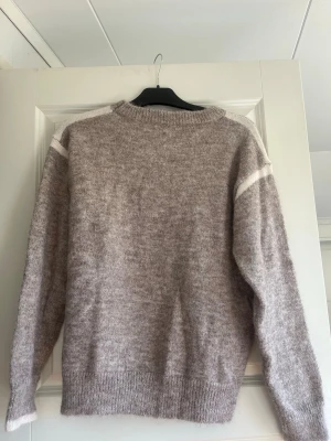 Beige stickad tröja med vit detalj - Mysig stickad tröja i beige med vita ränder på axlarna. Perfekt för dig som gillar en enkel och stilren look. Aldrig använd!