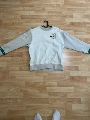 Vit Nike sweatshirt med gröna detaljer - Nike sweatshirt i vitt med grå muddar och gröna detaljer på ärmslut och hals. Broderad 'N' logga på bröstet. Klassisk rund hals och långärmad modell, perfekt för en avslappnad och sportig look.