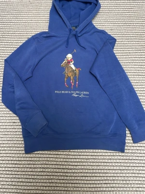 Blå hoodie Polo Bear Ralph Lauren - Blå hoodie från Ralph Lauren med Polo Bear-tryck på bröstet där björnen rider häst och har röd-svart tröja. Klassisk huva och ribbade muddar. Snygg streetwear vibe och perfekt för dig som gillar ikoniska prints och skön passform.