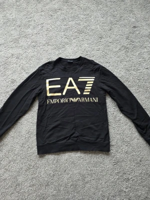 Svart EA7 Emporio Armani sweatshirt - Cool svart sweatshirt från EA7 Emporio Armani med stort guldfärgat tryck på bröstet. Tröjan har rund halsringning och långa ärmar. Perfekt för dig som gillar stilrena och exklusiva märken.
