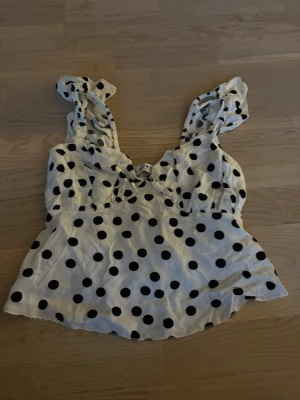 Prickig topp från Meet Me There - Supersöt vit topp med svarta prickar från Meet Me There. Toppen har breda axelband med volangdetaljer och en lätt utsvängd passform. Perfekt för dig som gillar lekfulla mönster och vill sticka ut lite extra.