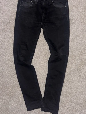 Svarta slim fit jeans från Nudie - Säljer ett par svarta slim fit jeans från Nudie med klassisk femficksdesign och diskreta sömmar. Jeansen har en modern passform och är tillverkade i mjukt denimtyg som sitter snyggt. Perfekta för dig som gillar stilrena och tidlösa plagg.