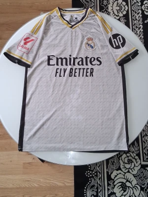 Real Madrid matchtröja Vini Jr. - Säljer en Real Madrid matchtröja med Vini Jr. och nummer 7 på ryggen. Tröjan är vit men de är dålig med ljus med svarta och gula detaljer, korta ärmar och klubbmärke på bröstet. Sponsorer som Emirates och La Liga-logga finns tryckta. Perfekt för fotbollsfans!