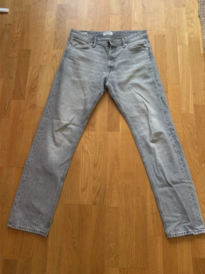 Grå raka jeans från Jack & Jones - Snygga grå jeans från Jack & Jones med rak passform och klassisk femficksdesign. Jeansen har en ljus tvätt och silverfärgade nitar. Perfekta för en avslappnad och trendig stil. Materialet är bomull med en mjuk känsla.
