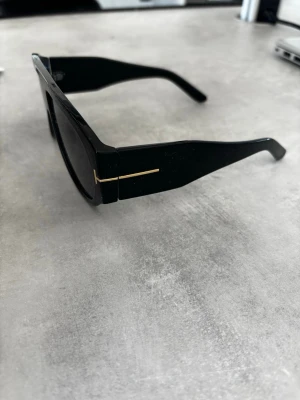 Tom Ford solglasögon - Stilrena svarta solglasögon med bred, markerad båge och diskret gulddetalj vid gångjärnet. Rektangulär form som ger en modern vibe och passar till de flesta outfits. Tillverkade i syntetmaterial för en lätt och bekväm känsla.