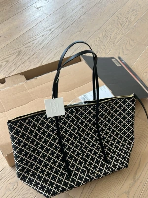 Svart axelväska By Malene Birger Luze  - Snygg svart axelväska från By Malene Birger med vitt grafiskt mönster och guldfärgad dragkedja. Väskan har långa svarta axelband och är tillverkad i tåligt material. Perfekt för dig som vill ha en trendig och rymlig väska med unik design.