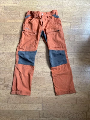 Orange byxa från Lundhags - Snygga orange byxor från Lundhags med grå förstärkningar på knän och fickor. Byxorna har dragkedjor på fickorna och knappstängning i midjan. Perfekta för dig som gillar funktionella och tåliga plagg.