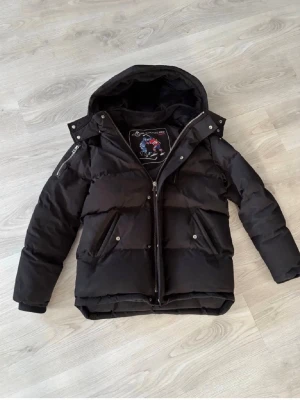 Svart pufferjacka med huva - Säljer en svart pufferjacka från Moose Knuckles med avtagbar huva och dragkedja framtill. Jackan har två snedställda fickor med tryckknappar och en patch på insidan. Perfekt för kalla vinterdagar och riktigt snygg streetstil.