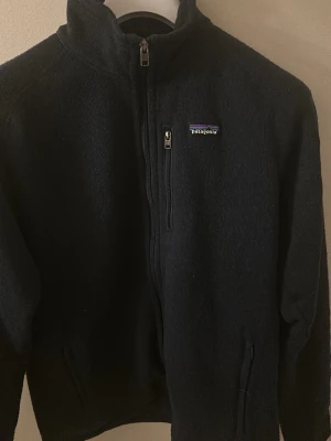 Mörkblå fleecejacka från Patagonia - Mysig blå fleecejacka från Patagonia med hög krage och dragkedja framtill. Jackan har två sidofickor med dragkedja och en bröstficka med logga. Perfekt för kyliga dagar och enkel att matcha till olika outfits. I princip som ny.