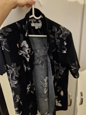 Svart blommig skjorta från SNDST - Snygg svart skjorta från SNDST med vita och grå blommor. Skjortan har korta ärmar, klassisk krage och knappar framtill. Perfekt för dig som vill sticka ut lite extra med ett coolt blommönster. Tillverkad i ett mjukt och lätt material.