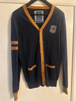 Mörkblå cardigan med gula detaljer - Stilren mörkblå cardigan inspirerad av Hogwarts med gula detaljer längs kanter, muddar och fickor. Två gula ränder på ena ärmen och Hogwarts-emblem på bröstet. Perfekt för dig som gillar magiska detaljer och college-stil.