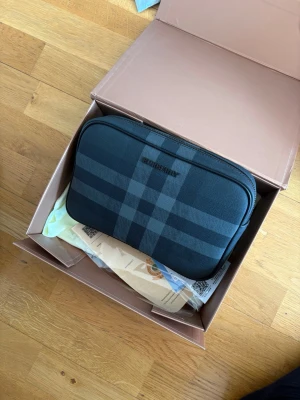 Svart rutig handväska från Burberry - Snygg svart handväska från Burberry med klassiskt rutmönster i mörkgrå toner. Väskan har dragkedja upptill och är tillverkad i canvas med detaljer i läder. Perfekt för dig som gillar stilrena accessoarer med lyxig känsla.