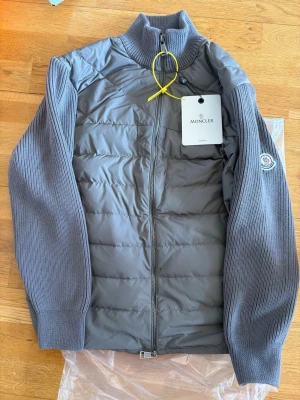 Grå cardigan från Moncler - Snygg grå pufferjacka från Moncler med stickade ärmar och hög krage. Jackan har quiltad front, två sidofickor och Moncler-logga på ärmen. Materialet är en kombination av dun och stickad textil, perfekt för kalla vinterdagar.