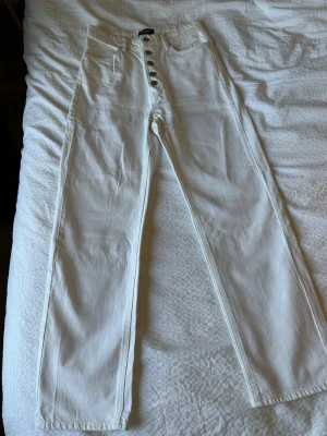 Vita raka jeans med knappgylf - Snygga vita jeans från Lindex med raka ben och hög midja. Jeansen har knäppning med flera silverfärgade knappar framtill. Ankellängd se bild 4. Endast använda 2/3 gånger. Jag är 168cm