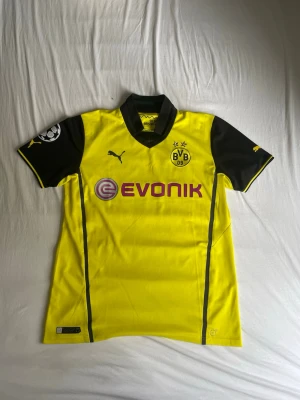 Borussia Dortmund matchtröja Puma - Gul och svart Borussia Dortmund matchtröja från Puma med tryck 'Mkhitaryan 10' på ryggen. Tröjan har klubbmärke, sponsorlogga och Champions League-märke på ärmen. Klassisk krage och korta ärmar, tillverkad i funktionsmaterial.