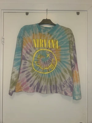Nirvana tie-dye långärmad tröja - Färgglad långärmad tröja med tie-dye mönster i blått, gult, lila, grönt och orange. Stort gult Nirvana-tryck och smiley på bröstet. Perfekt för dig som gillar bandtröjor och retrostil. Väldigt stor och oversized passar mig som har L