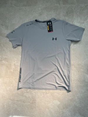 Ljusgrå träningströja Under Armour - Snygg ljusgrå t-shirt från Under Armour med svart logga på bröstet och tryck på ärmen samt längs sidan. Tillverkad i ett lätt och snabbtorkande material, perfekt för träning och sportaktiviteter. Helt ny med prislapp! Storlek: S. Pris går att diskutera☺️